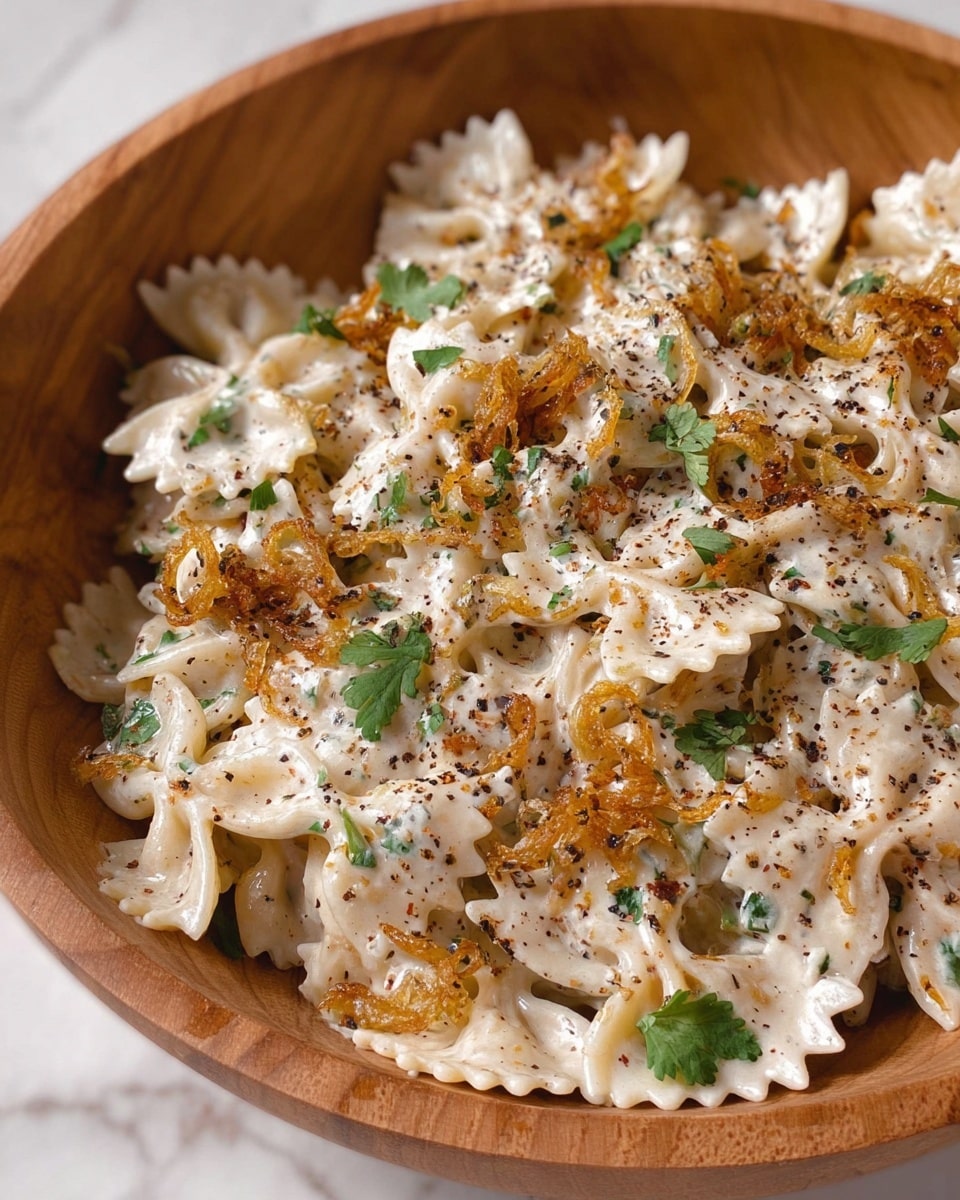 The dish shows a wooden bowl filled with creamy white farfalle pasta layered evenly throughout. The creamy sauce covers every piece, speckled with tiny green herbs adding texture. On top, there are golden brown crispy fried onion slices scattered with some fresh green cilantro leaves for color contrast. The light coating of black pepper is visible over the whole dish, making it look flavorful and rich. The bowl is placed on a white marbled surface. photo taken with an iphone --ar 4:5 --v 7