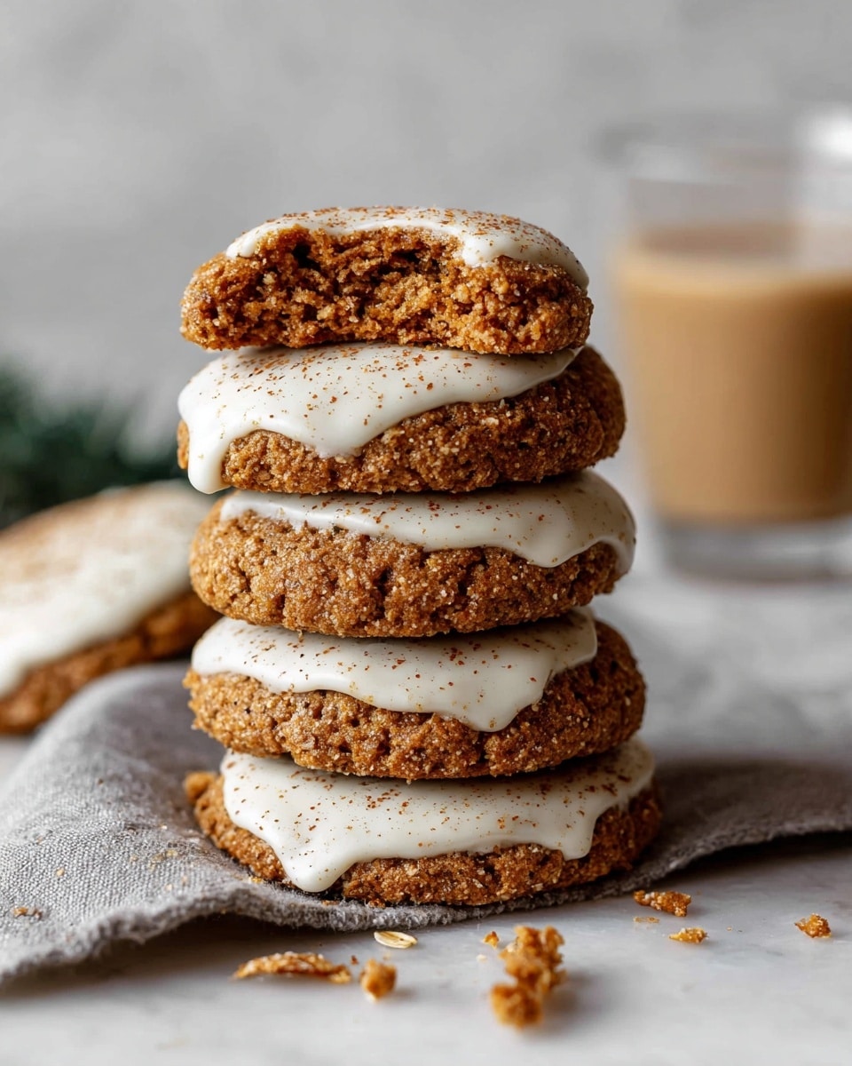 A stack of five round cookies with a rough, crumbly texture sits on a light gray cloth over a white marbled surface. Each cookie is partially dipped in smooth, creamy white icing sprinkled with fine brown specks, covering about one-third of each cookie on the side. The top cookie is broken in half, showing a dense, slightly chewy inside with bits of oats or nuts visible. Small crumbs are scattered around the stack, and in the background, there is a blurred clear glass filled with light brown milk or coffee. photo taken with an iphone --ar 4:5 --v 7