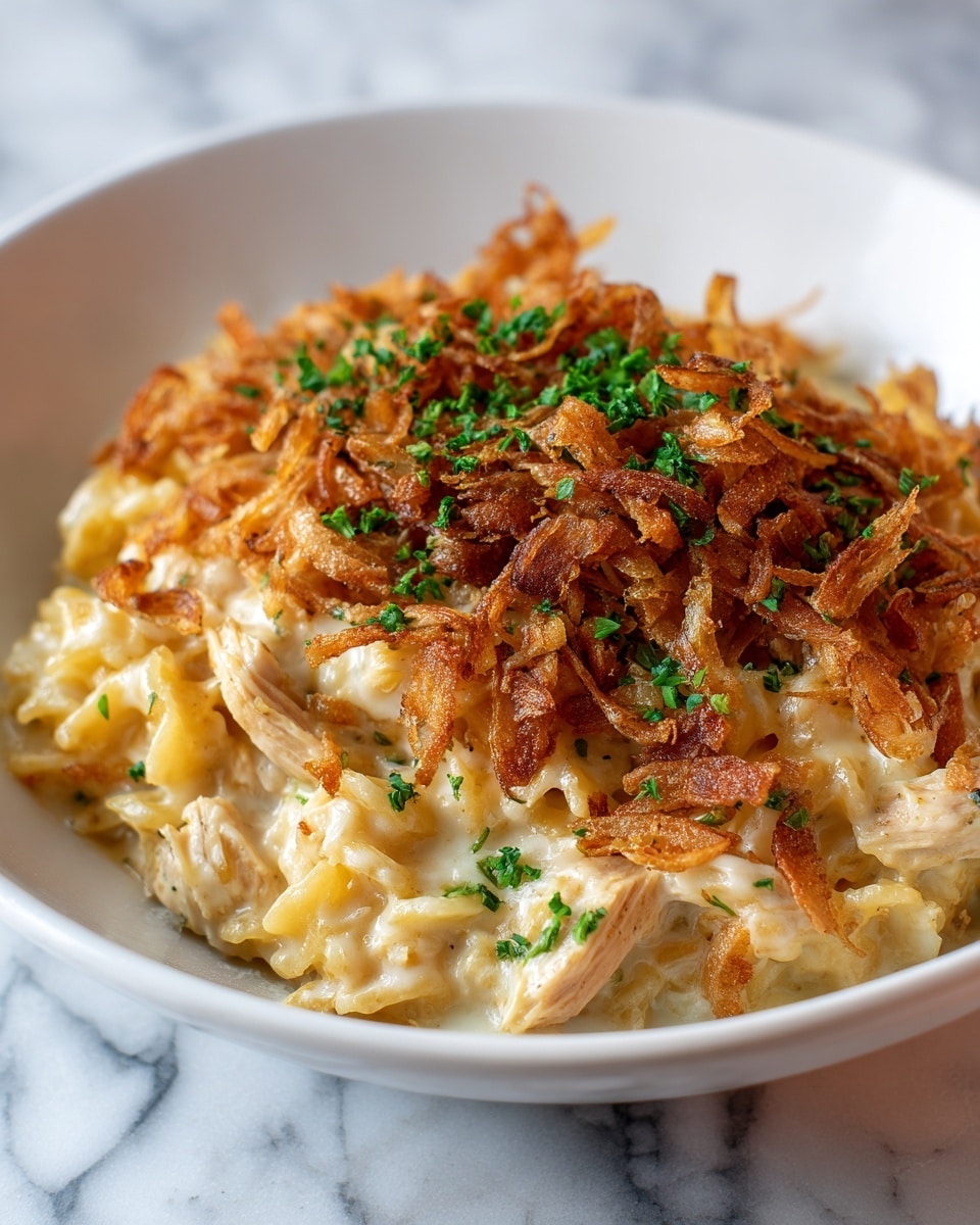 The dish is a creamy casserole served in a white bowl placed on a white marbled surface. The bottom layer consists of a soft, cheesy pasta, pale yellow and smooth in texture. Mixed and slightly buried in the pasta are tender pieces of light golden brown chicken. On top of the pasta and chicken is a crispy brown layer of fried onions with a rough, crunchy texture. Bright green chopped herbs are sprinkled evenly over the whole dish, adding a fresh contrast to the warm colors below. Photo taken with an iphone --ar 4:5 --v 7