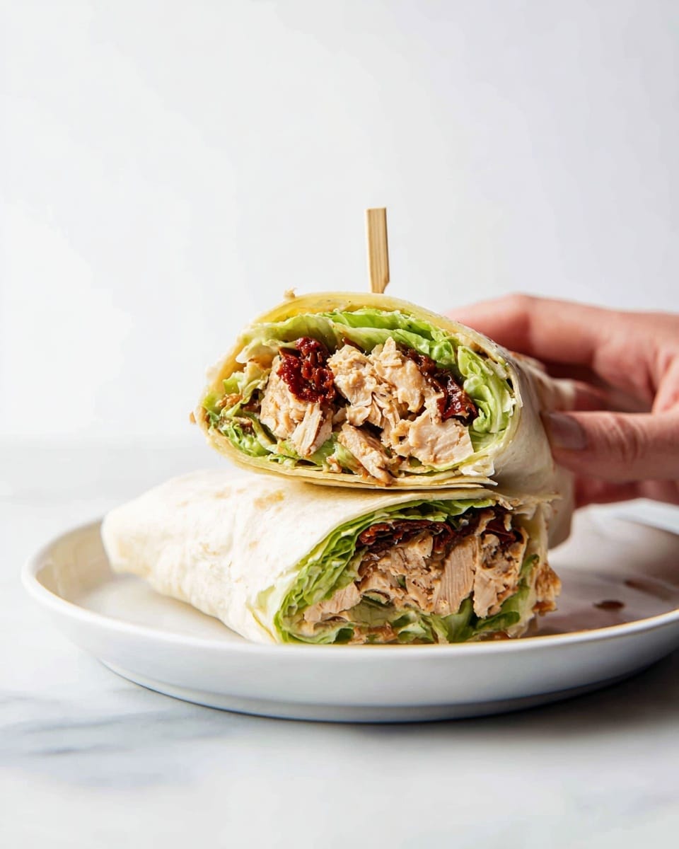 A wrap cut in half and stacked on a white plate with a white marbled background, showing three main layers inside: the outer soft light beige tortilla, a middle layer of green lettuce with a fresh texture, and a thick inner filling made of light brown chicken mixed with small pieces of dark red sun-dried tomatoes, all held together by a wooden skewer. A woman's hand is gently holding the edge of the plate, focusing on the wrap. photo taken with an iphone --ar 4:5 --v 7