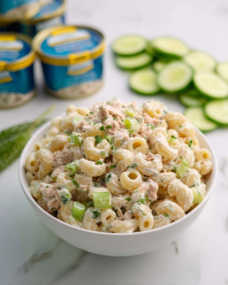 A white bowl filled with creamy macaroni salad sits on a white marbled surface. The salad has small curled pasta shells mixed with light beige tuna pieces and chopped light green celery, all coated in a smooth, slightly off-white dressing. Small bits of green herbs and scallions are scattered throughout the mixture, adding subtle color contrast. In the background, sliced cucumber rounds and several small canned tuna containers with blue and yellow lids are lightly blurred. Photo taken with an iphone --ar 4:5 --v 7