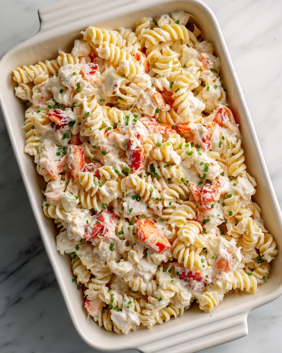 A white rectangular baking dish filled with a creamy pasta salad made of three main layers: soft, pale yellow twisted egg noodles mixed thoroughly with large chunks of white seafood meat, likely crab, and small pieces of bright red crab sticks scattered evenly throughout. The entire pasta is coated in a thick, white creamy dressing that binds all ingredients together, and small flecks of fresh green herbs or scallions are visible sprinkled across the dish, adding a touch of color contrast. The dish is placed on a smooth white marbled surface. photo taken with an iphone --ar 4:5 --v 7