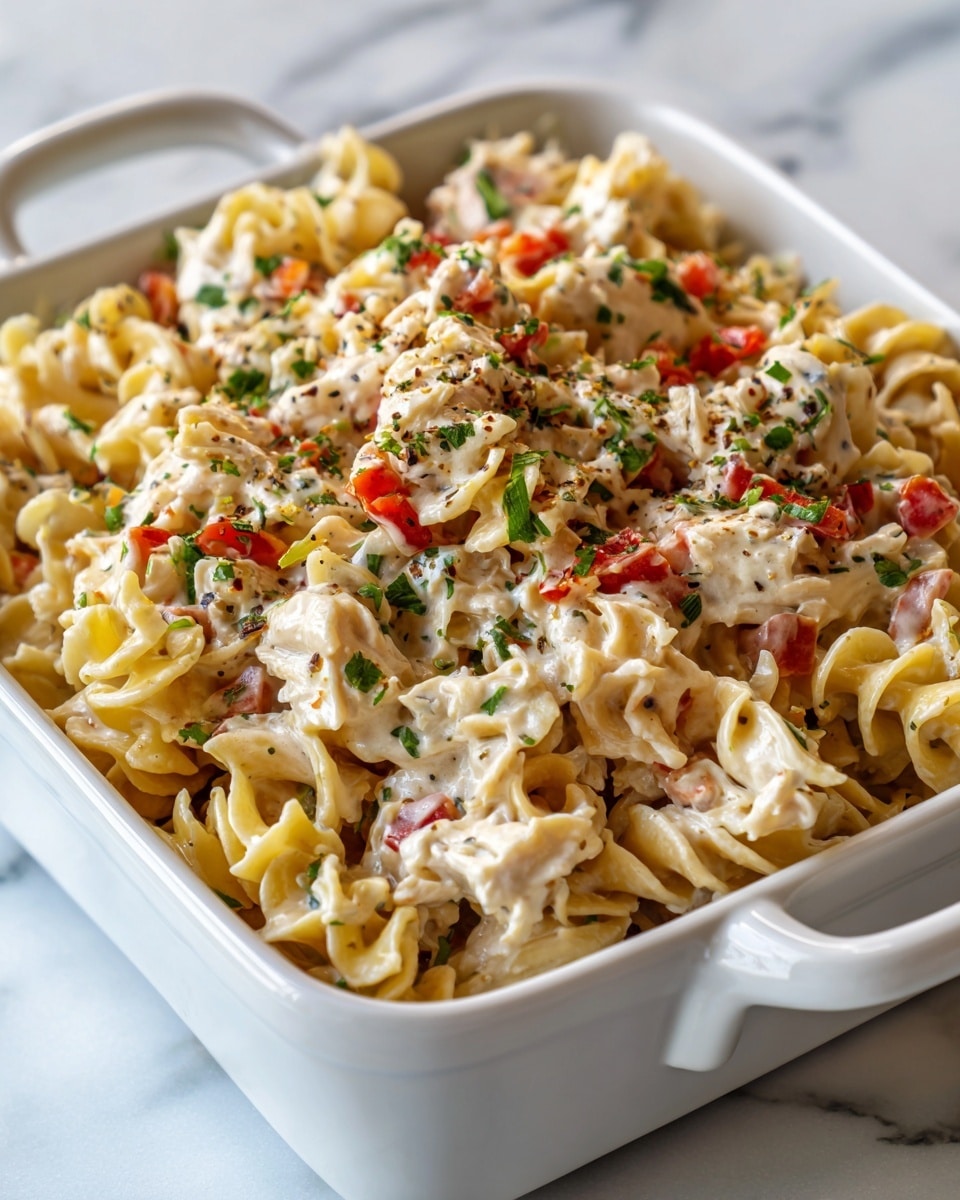 A creamy pasta salad sits in a white rectangular dish with handles on each side, placed on a white marbled surface. The salad has one visible layer composed of curly, yellow egg noodles coated in a thick white dressing mixed evenly with small chopped pieces of red bell pepper and bits of green herbs, as well as chunks of white chicken. The texture looks rich and smooth with a mix of soft pasta and tender chicken pieces throughout. Photo taken with an iphone --ar 4:5 --v 7