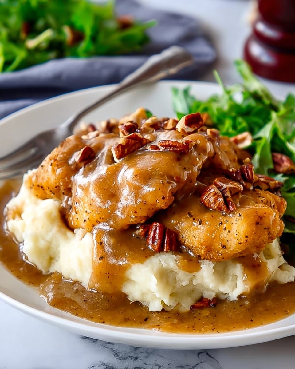 A white plate on a white marbled surface holds a base layer of creamy white mashed potatoes with a smooth texture. On top, there are three pieces of golden brown fried chicken covered in a thick, rich brown gravy with visible specks of seasoning. Scattered over the chicken and gravy are small, uneven pieces of chopped pecans adding a crunchy texture. In the background, there is a hint of fresh green leafy vegetables adding color contrast. A silver fork is placed near the plate. Photo taken with an iphone --ar 4:5 --v 7