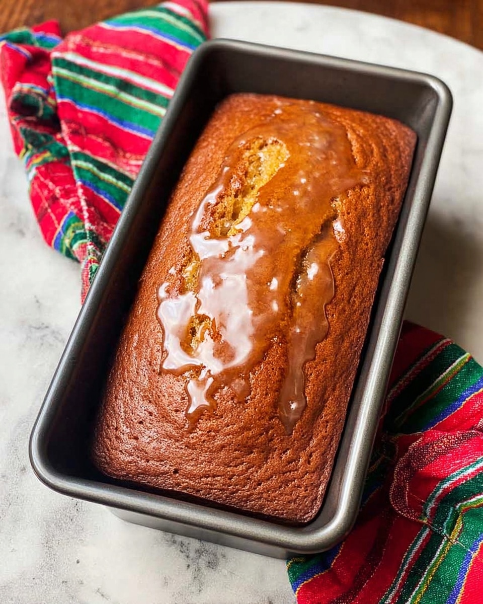 A single loaf of golden brown cake with a shiny glaze on top is placed inside a dark gray metal rectangular baking pan. The glaze pools slightly at the edges, giving the cake a moist look. The baking pan rests on a white marbled textured surface with a colorful red, green, and blue striped cloth casually draped around the bottom left corner of the pan. Photo taken with an iphone --ar 4:5 --v 7