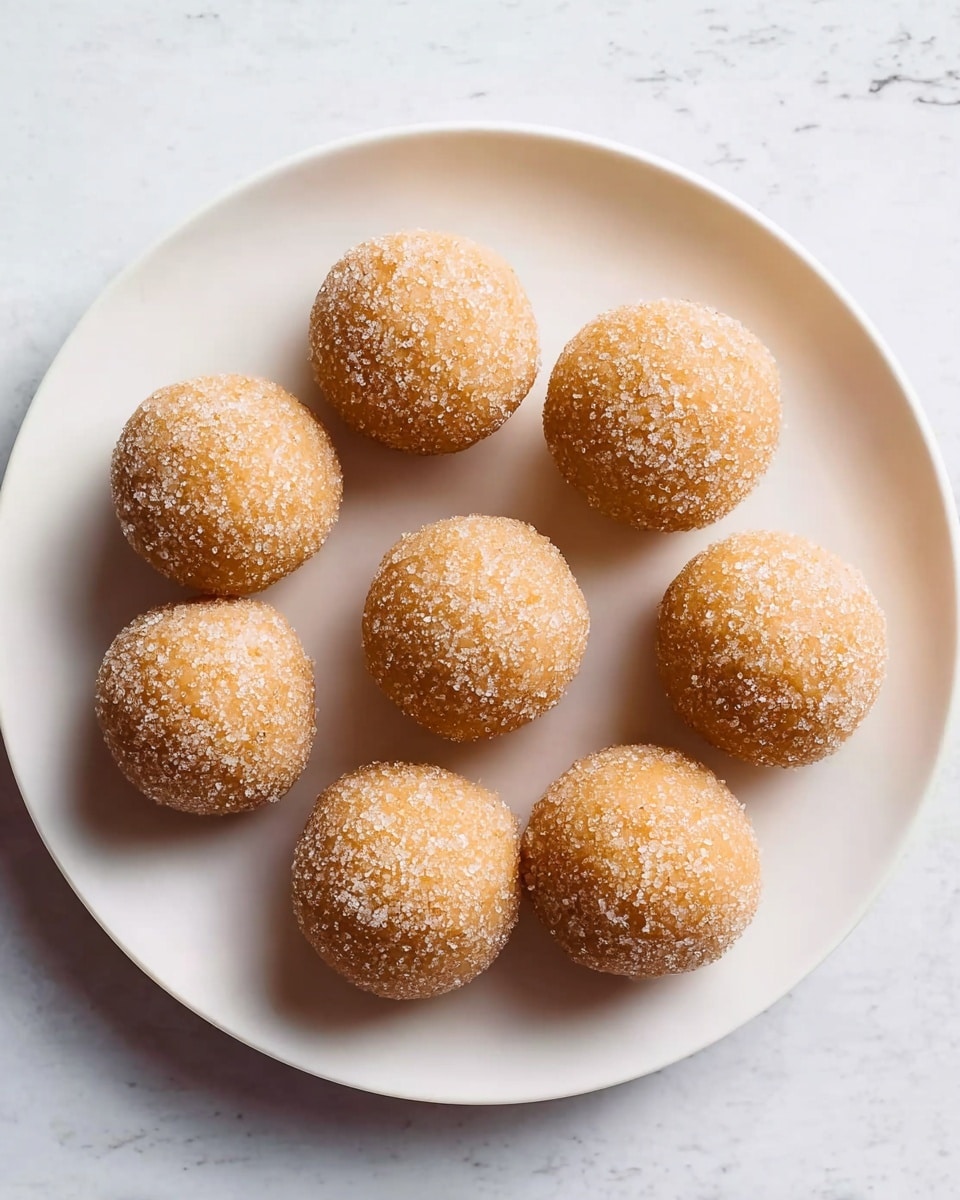 Nine round balls covered in sugar crystals are placed on a white plate arranged in a circular pattern with one ball in the center and eight around it. The balls have a light golden brown color and a slightly grainy texture from the sugar coating. The white plate sits on a white marbled textured surface, and the lighting highlights the sugar crystals making them sparkle slightly. photo taken with an iphone --ar 4:5 --v 7