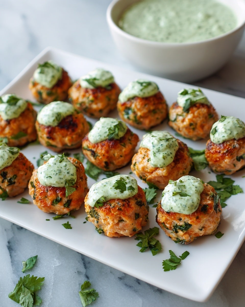 A white square plate holds twelve round salmon meatballs arranged in several rows; each meatball is light orange with green herbs mixed in, and they have a browned, slightly crispy texture on the outside. Many meatballs are topped with a dollop of creamy pale green sauce that looks smooth and thick, with small green flecks. Fresh chopped herbs are scattered around the meatballs on the plate, adding vibrant green accents. In the background, there is a white bowl filled with more of the same pale green sauce. The plate sits on a white marbled surface. photo taken with an iphone --ar 4:5 --v 7