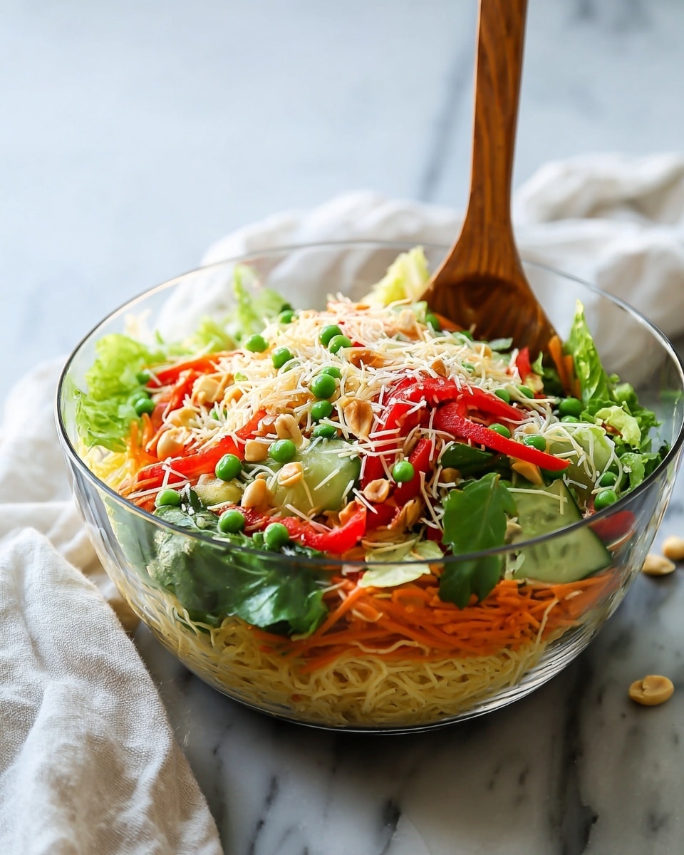 A clear glass bowl holds a colorful layered salad set on a white marbled surface with a soft white cloth nearby. The bottom layer consists of thin, light yellow noodles mixed with some shredded orange carrot. On top of this rests a mix of fresh green leafy lettuce, green peas, bright red bell pepper strips, and sliced cucumber with light green edges and white centers. The salad is sprinkled with shredded white cheese and chopped nuts, adding texture and contrast. A wooden spoon stands upright inside the bowl, slightly tilted towards the front. Photo taken with an iphone --ar 4:5 --v 7
