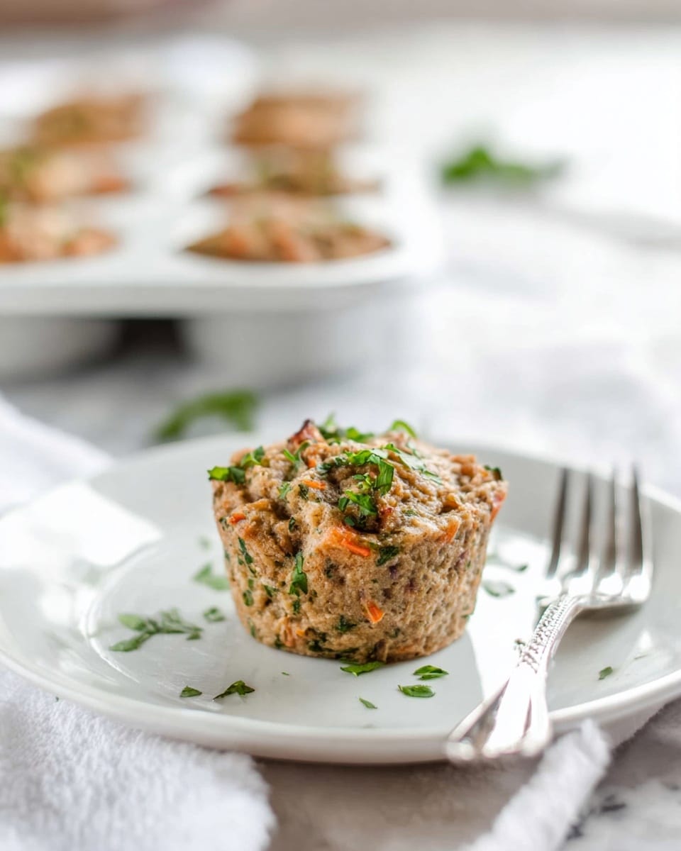 The image shows a single savory muffin on a white plate placed on a white marbled surface. The muffin has a rough texture with visible bits of green herbs and small orange pieces scattered throughout its light brown body. It is garnished with small chopped green herbs on top and around the plate. In the background, there is a white muffin tin holding several more muffins, slightly out of focus. A silver fork rests to the right side of the plate. photo taken with an iphone --ar 4:5 --v 7