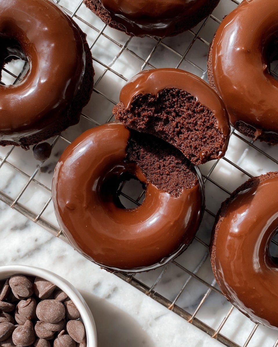 The image shows several chocolate-glazed donuts cooling on a metal rack over a white marbled surface. The donuts have a thick, shiny layer of smooth, dark brown chocolate glaze covering the entire top, creating a glossy finish. One donut is in the center with a bite taken out, revealing a rich, dark, moist chocolate cake inside with a soft texture. In the bottom left corner, there is a white bowl filled with small, round milk chocolate chips. The lighting highlights the gloss on the glaze and the texture of the cake inside the bitten donut. photo taken with an iphone --ar 4:5 --v 7