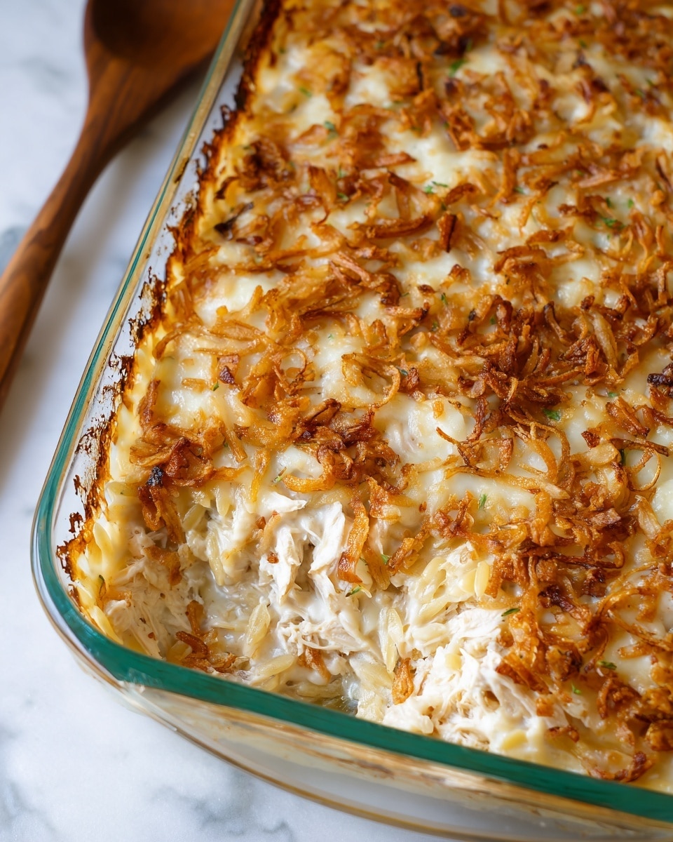 The image shows a close-up of a baked dish in a clear glass rectangular baking dish with green edges. The dish has a creamy textured layer of melted white cheese on top, sprinkled unevenly with golden brown crispy fried onions that add a crunchy texture. Below the cheese top layer, visible near the edge, are soft, cooked, pale yellow orzo pasta pieces mixed with shredded white meat, possibly chicken. The edges of the baking dish have some browned baked residue, and the dish sits on a white marbled textured surface. A wooden spoon is partially visible on the left in the background. Photo taken with an iphone --ar 4:5 --v 7