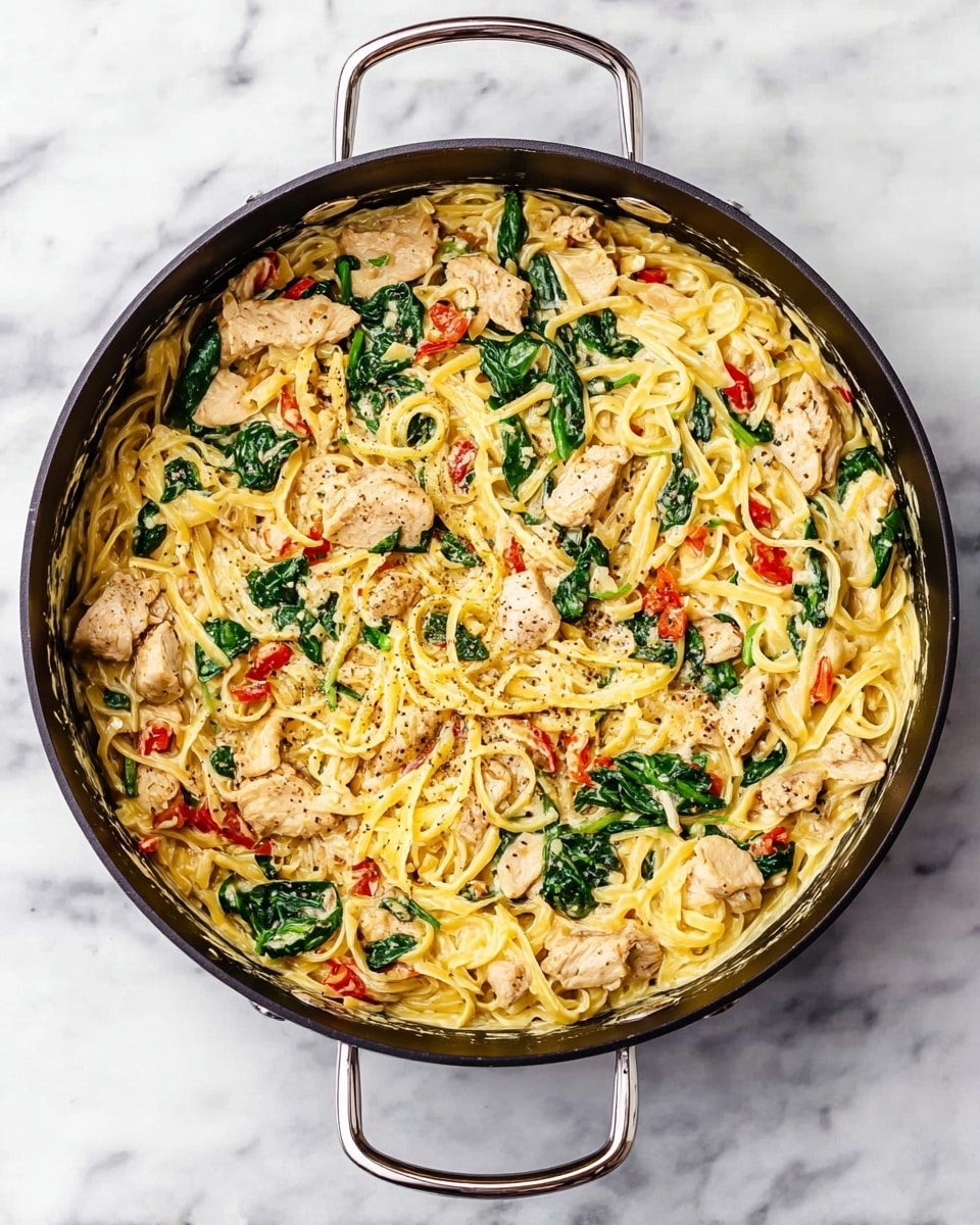 A top-down view of a large black pan filled with a creamy pasta dish. The dish has one main layer with creamy yellow pasta mixed with pieces of cooked chicken, bright green spinach leaves, and small red roasted pepper strips all evenly spread. The chicken pieces are light brown with visible seasoning, while the spinach and red peppers add fresh color contrast. The pan has silver handles and is placed on a white marbled surface. photo taken with an iphone --ar 4:5 --v 7