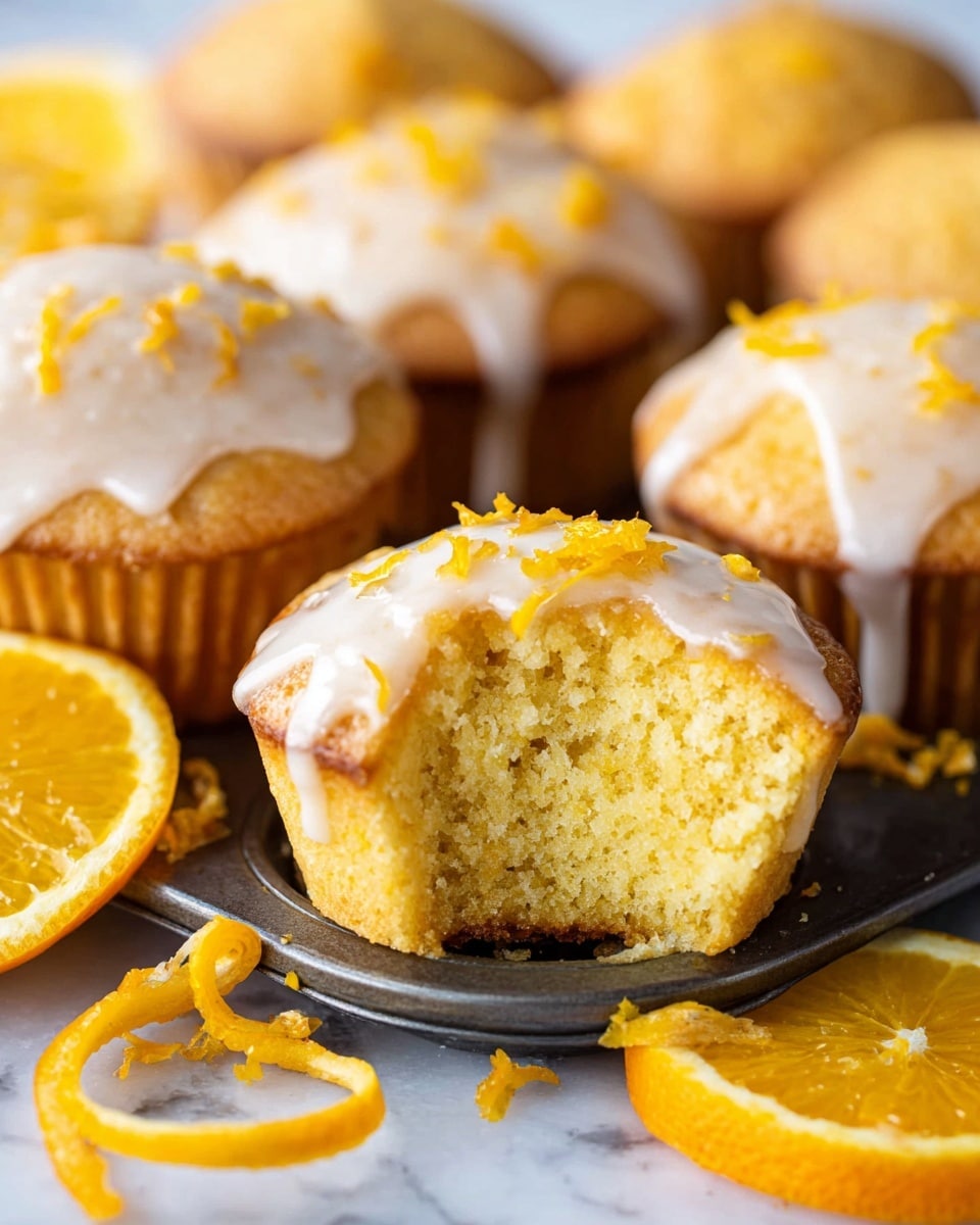 The image shows a close-up of several orange-glazed muffins in a muffin tray with a white marbled surface beneath. Each muffin has one layer of soft, golden yellow cake topped with a shiny white glaze that drips slightly down the sides. One muffin is bitten, revealing a moist, crumbly inside with a pale yellow color. Bright orange zest is sprinkled on top of the glaze, adding texture and color contrast. Around the muffins, there are thin, curly orange peels and slices of fresh orange, creating a fresh, bright look. Photo taken with an iphone --ar 4:5 --v 7