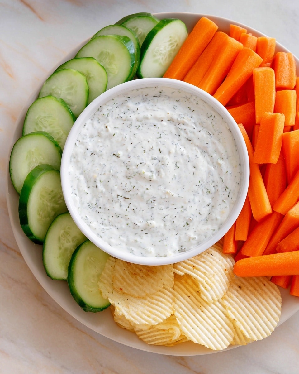 A white bowl filled with creamy white dip flecked with small green herbs is sitting in the middle of a large white plate. Around the bowl, there is a ring of bright orange baby carrots on the right, fresh green cucumber spears lined up neatly on the bottom and left, and a few pale yellow ridged potato chips placed near the carrots. The whole plate is placed on a white marbled surface. photo taken with an iphone --ar 4:5 --v 7