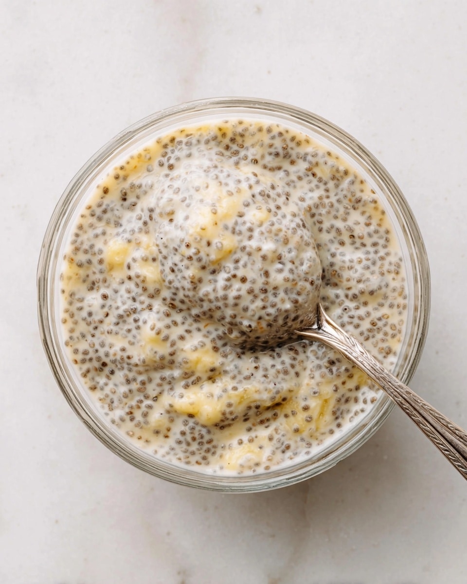 A clear glass bowl filled with a creamy, light beige chia pudding mixed with small chunks of soft yellow banana throughout, creating a speckled and slightly lumpy texture. A silver spoon scoops a portion of the pudding from the center, showing the thick consistency and tiny black chia seeds suspended evenly in the mixture. The bowl sits on a white marbled surface, giving a clean and simple background. photo taken with an iphone --ar 4:5 --v 7