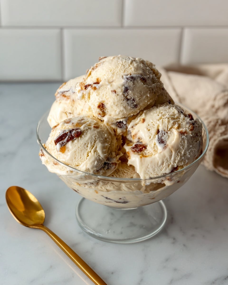 A clear glass bowl filled with five scoops of creamy, light beige ice cream mixed with dark brown and amber chunks all throughout, resting on a smooth white marbled surface. To the left of the bowl lies a shiny gold spoon with a slightly curved handle. The background is a white tiled wall with a soft, neutral-colored cloth partially visible on the right side. The ice cream has a soft, creamy texture with well-distributed pieces giving it a textured look. photo taken with an iphone --ar 4:5 --v 7