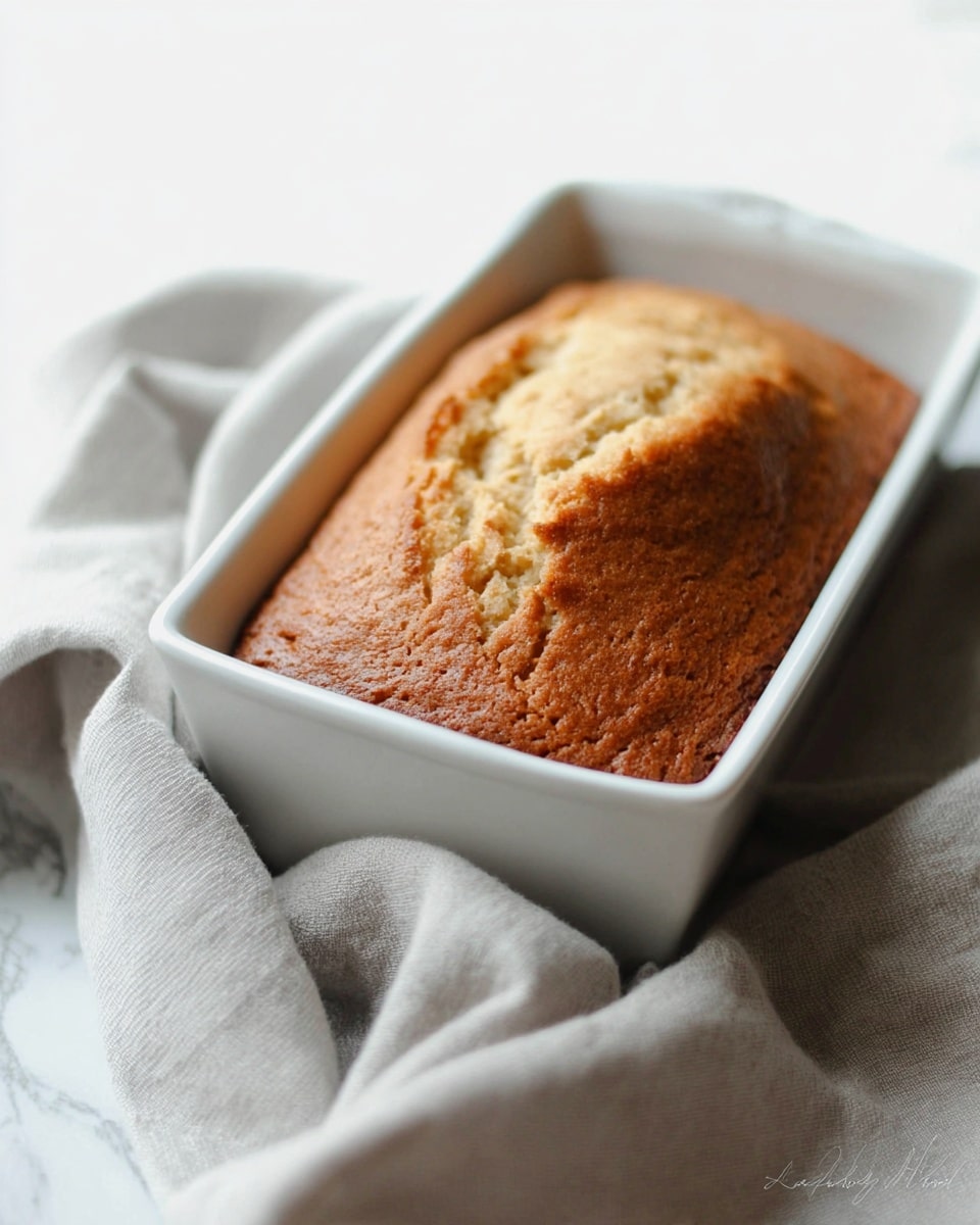 A golden brown loaf cake with a slightly cracked and textured top layer fills a white rectangular baking dish, which is sitting on a soft, light gray cloth draped over a white marbled surface. The cake's top has a warm, fluffy appearance with light air pockets visible, showing a fresh, baked texture. The overall scene has a simple, cozy feel and soft natural lighting. photo taken with an iphone --ar 4:5 --v 7