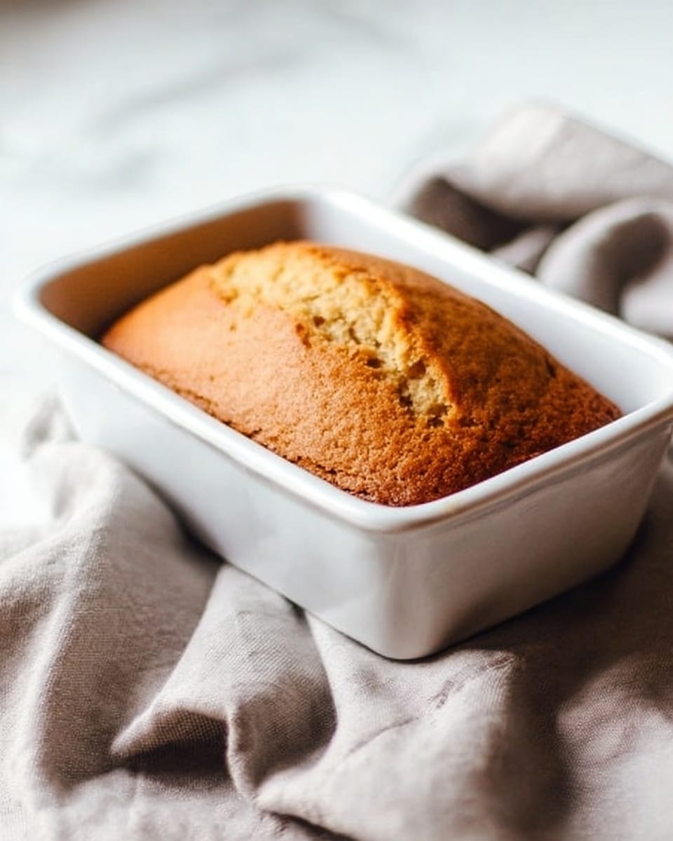 A golden brown loaf cake with a cracked top is inside a small white rectangular baking dish. The cake looks soft and moist with a spongy texture. The baking dish sits on a crumpled light gray cloth on a white marbled surface, softly lit from the left side. The scene is simple and clean, focusing on the cake’s warm color and the white dish. photo taken with an iphone --ar 4:5 --v 7