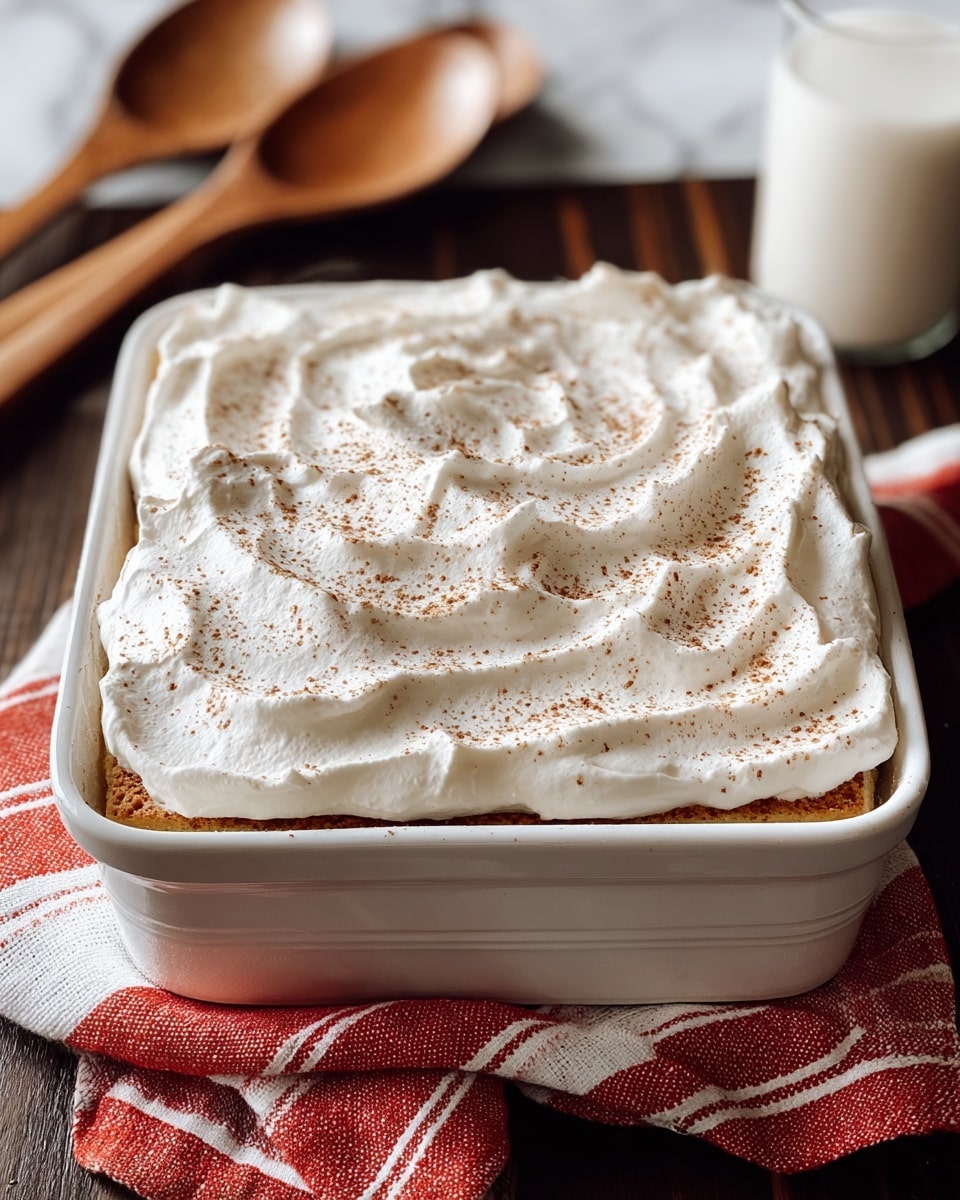 A square white ceramic dish holds a layered dessert topped with a thick, fluffy layer of white whipped cream spread unevenly with visible soft peaks and swirls, lightly sprinkled with a fine dusting of brown spice, likely cinnamon or nutmeg. The layers underneath the whipped cream are slightly visible at the edges, showing a light golden brown cake base. The dish is placed on a red and white striped cloth on a dark wooden table with a soft white marbled texture background, next to two wooden spoons and a partially visible glass of milk. photo taken with an iphone --ar 4:5 --v 7