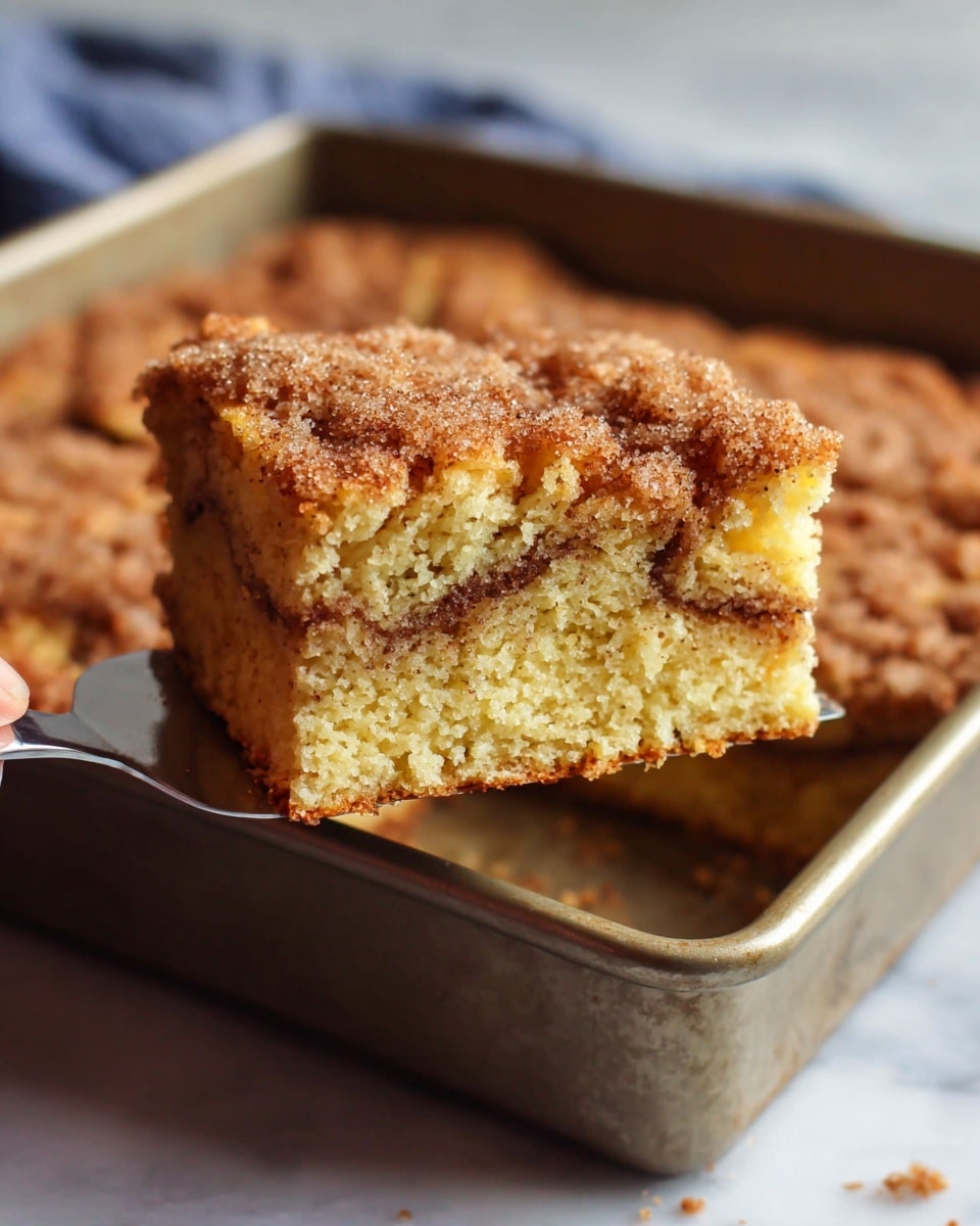 A close-up view of a single square slice of cinnamon coffee cake being lifted from a square baking pan, showing two visible layers: a light golden yellow moist cake base with a visible swirl of darker cinnamon filling inside, topped with a crumbly, textured brown sugar and cinnamon streusel layer. The slice has a rough but soft edge, and the streusel topping appears slightly crunchy with a sprinkle of sugar crystals. The background has a white marbled texture beneath the pan, and woman's hand is holding the spatula under the slice. Photo taken with an iphone --ar 4:5 --v 7
