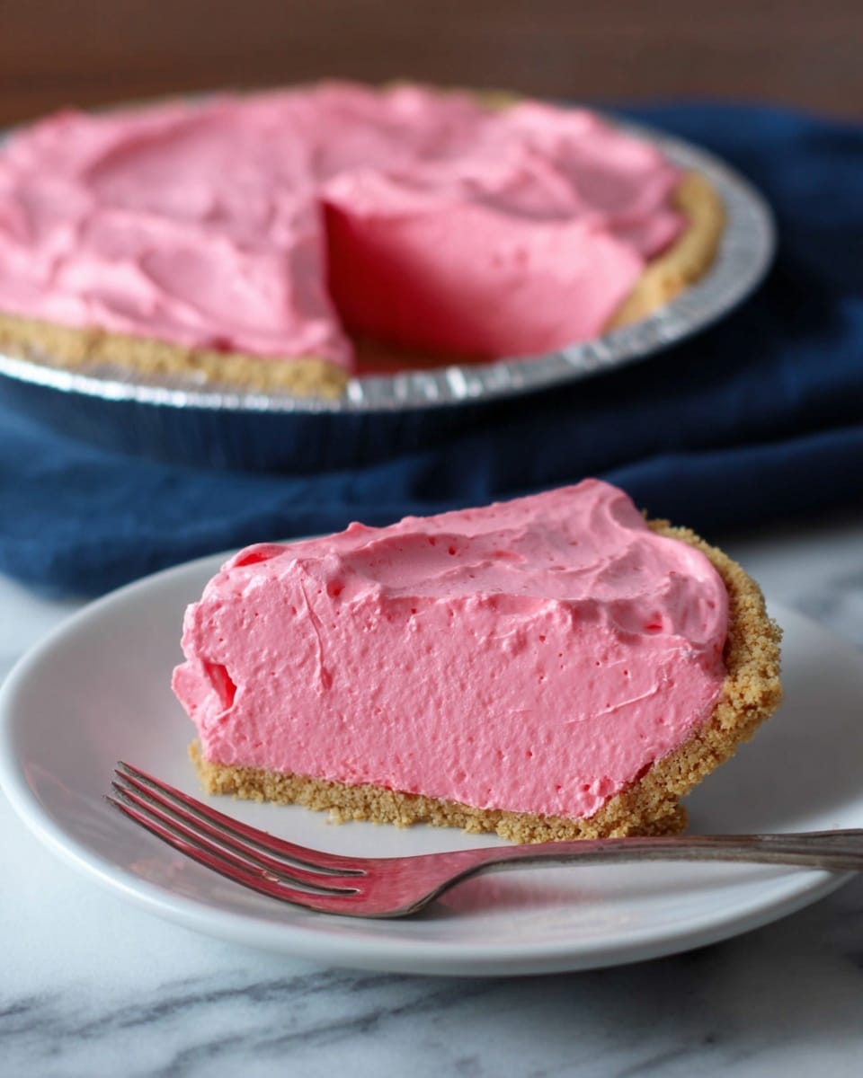 A single slice of bright pink creamy pie sits on a white plate in the foreground, showing three layers: a rough, light brown crumbly crust at the bottom, a thick, smooth, and airy bright pink filling in the middle, and a slightly denser pink topping spread evenly on top. The slice is accompanied by a shiny fork resting on the plate's edge. In the background, the rest of the pie with the same three layers is in a silver foil pie pan, with one slice already removed, placed on a white marbled texture surface partially covered by a dark blue cloth. The lighting is soft and natural. photo taken with an iphone --ar 4:5 --v 7