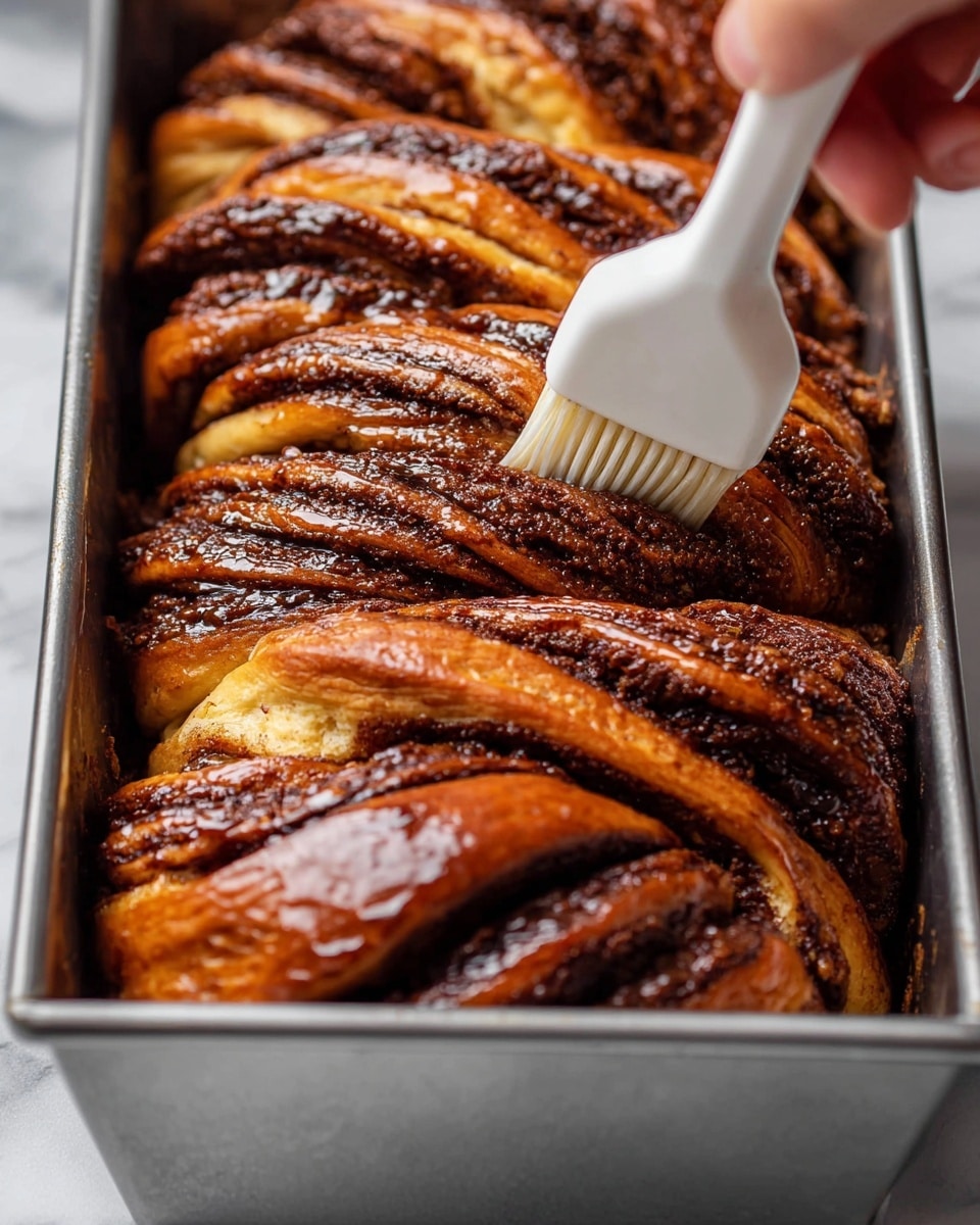 A close-up view of a large baked cinnamon babka inside a metal baking pan, showing multiple twisted layers of golden brown dough with dark brown cinnamon filling between them, glistening with a shiny glaze. A woman's hand is brushing the glossy surface of the babka with a white silicone brush, emphasizing the rich texture and soft, fluffy bread layers in between the dense cinnamon swirls. The image is set against a white marbled surface. photo taken with an iphone --ar 4:5 --v 7