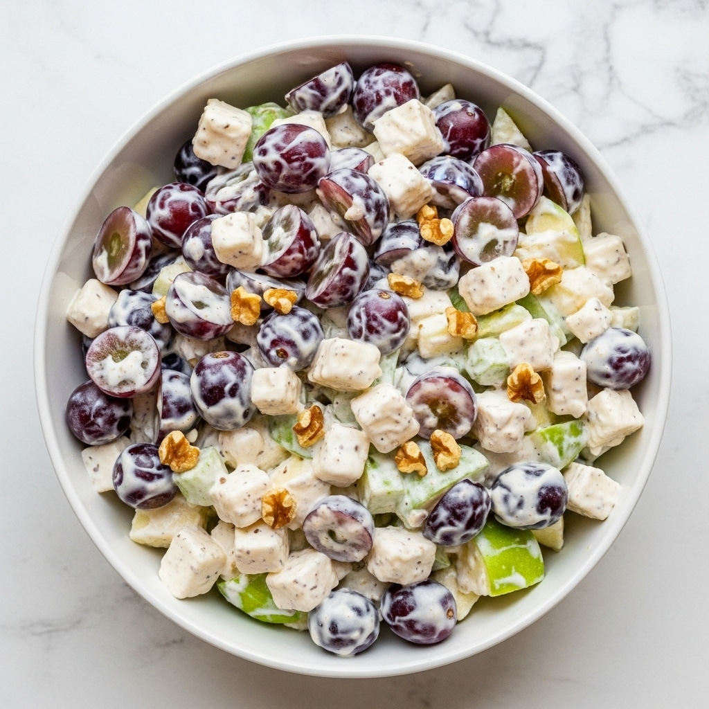 A white bowl filled with a creamy fruit salad sits on a white marbled texture. The salad has three main layers: purple grape slices, small white cubes of cheese, and green apple pieces, all mixed with a thick, white creamy dressing that coats the fruit and cheese evenly. Small bits of walnut are sprinkled on top and inside the salad, adding a crunchy texture. The bowl is full and the fruit pieces are clearly visible, showing their juicy and fresh qualities. Photo taken with an iphone --ar 4:5 --v 7