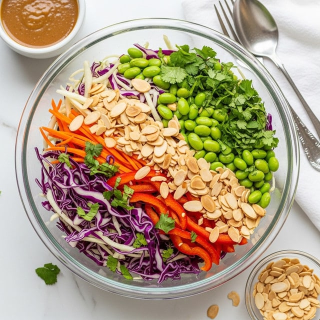 A clear glass bowl filled with a colorful mixed salad sits on a white marbled surface. The salad has three main layers: a base of thin white and purple cabbage strips, mixed with bright orange carrot sticks and red bell pepper slices, topped with scattered green edamame beans and sprinkled with fresh chopped green herbs, likely cilantro. Thin, toasted almond slices are spread evenly across the top, adding a light beige color and textural contrast. In the background to the top left, there is a small white bowl with brown dressing, and to the bottom right, a small clear bowl filled with extra toasted almond slices. A silver fork and spoon rest near the bowl on a white cloth. photo taken with an iphone --ar 4:5 --v 7