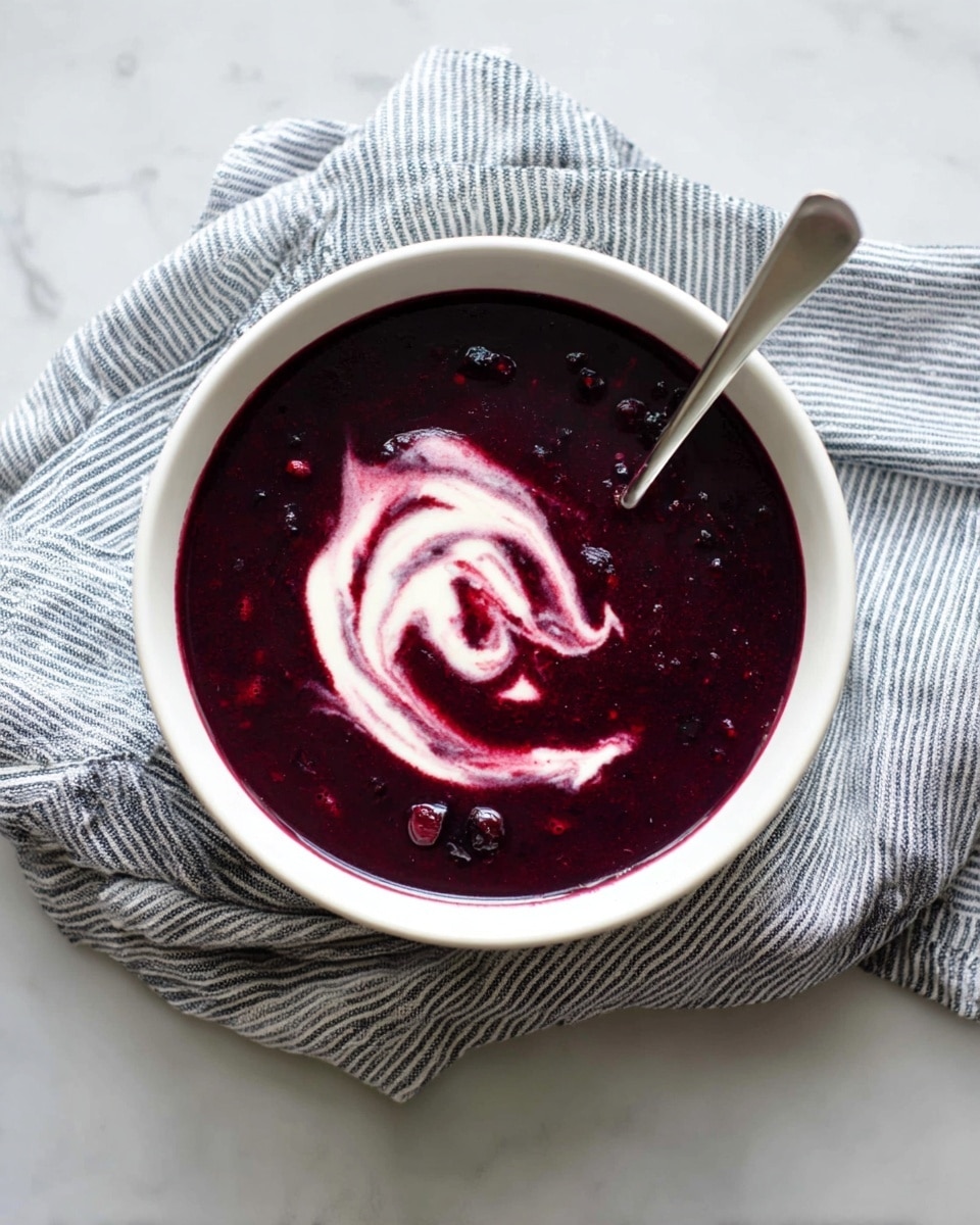 A white bowl filled with a dark purple soup that looks thick and smooth with some small berry pieces visible. In the center, there is a swirl of white cream mixed softly into the soup, creating a marbled effect. A silver spoon rests inside the bowl on the right side, slightly immersed in the soup. The bowl sits on a gray and white striped cloth with a white marbled surface underneath. photo taken with an iphone --ar 4:5 --v 7