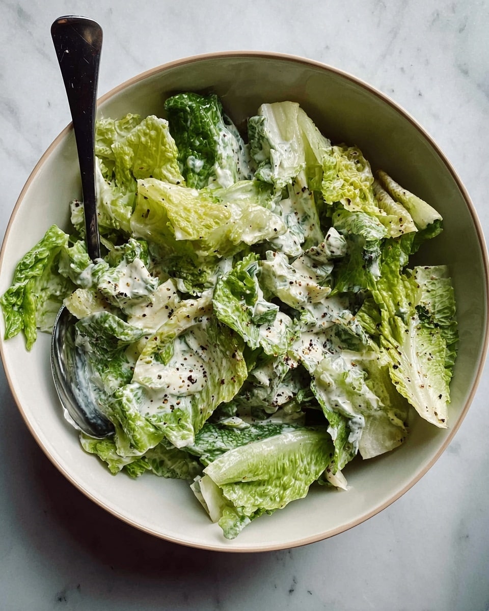A fresh salad is shown in a white bowl filled with large, crisp green lettuce leaves in different shades, some light green and others darker, covered with a creamy white dressing that holds small bits of herbs and spices. Scattered on top are tiny black seeds or spices, adding texture and contrast. The salad looks lightly mixed, with the leaves partially coated in dressing, and a silver fork with a black handle lies inside the bowl on the left side. The bowl is placed on a white marbled surface. photo taken with an iphone --ar 4:5 --v 7